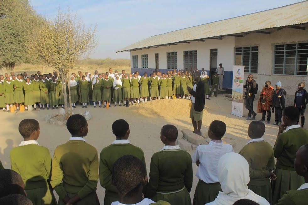 GACS speaker addressing students in green uniforms at a rural school courtyard
